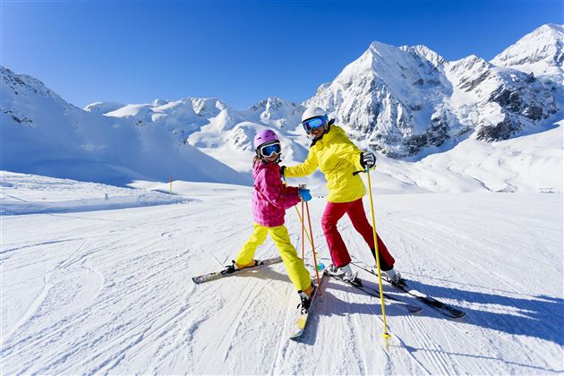 Zwei Kinder in bunten Skianzügen stehen auf einer schneebedeckten Skipiste. Im Hintergrund sind majestätische Berge unter einem klaren blauen Himmel zu sehen.