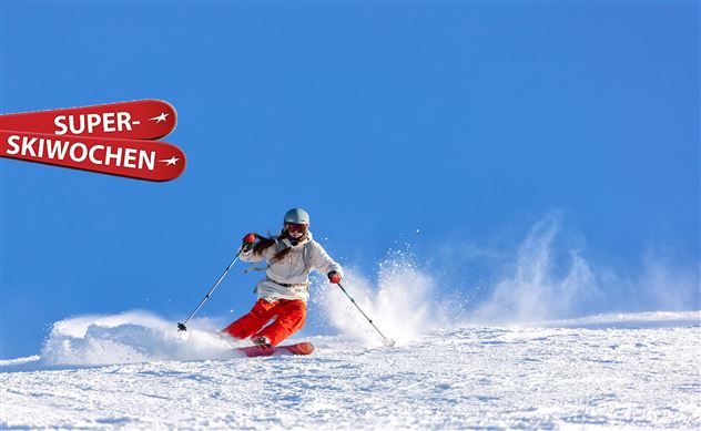 A person is skiing on a snow-covered slope under a clear blue sky. In the background, there is a sign that reads "SUPER SKI WEEKS."