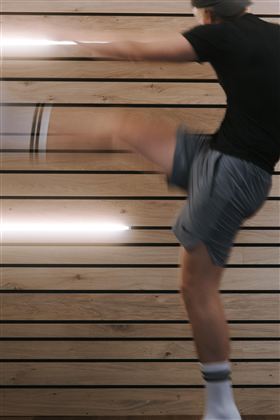 A man performs a dynamic kick. In the background, wooden-paneled walls and modern lighting are visible.