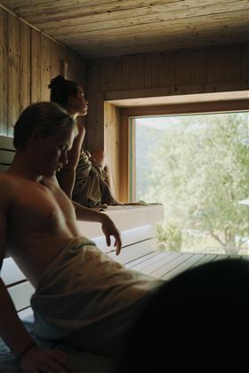 Two people are relaxing in a sauna. The room is made of wood and offers a view of the nature outside.