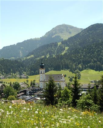 A picturesque mountain landscape with a small church and traditional buildings. In the foreground, meadow flowers are blooming, while the green hills gently rise in the background.