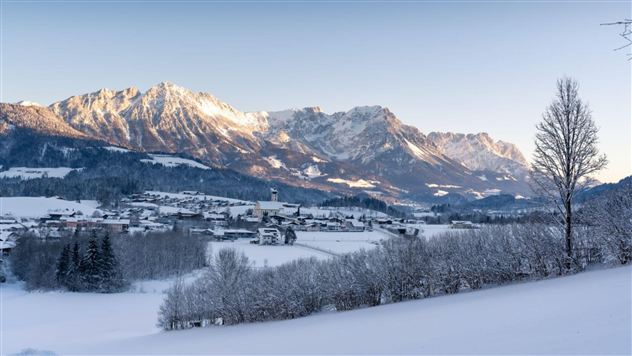 Eine winterliche Landschaft mit schneebedeckten Bergen und einem kleinen Dorf im Tal. Die Landschaft wird von sanften Lichtstrahlen der tiefstehenden Sonne beleuchtet.