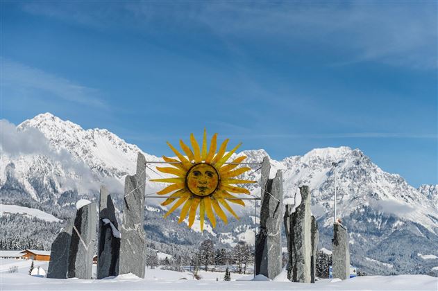 An impressive winter landscape with a sun sculpture and stone monuments. In the background, snow-covered mountains and a clear blue sky can be seen.