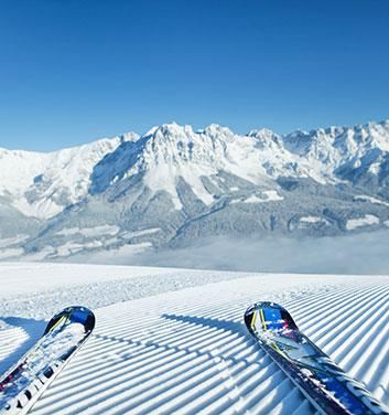 A snow-covered mountain landscape with a clear blue sky. In the foreground, two skis are lying on the groomed slope.