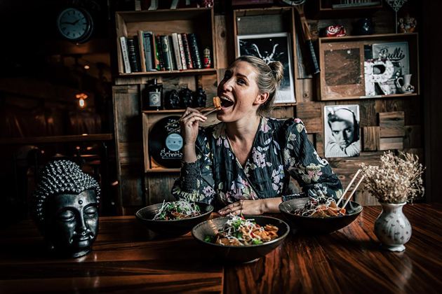 A woman is sitting at a table and enjoying Asian food. In the background, books and decorative elements are visible.