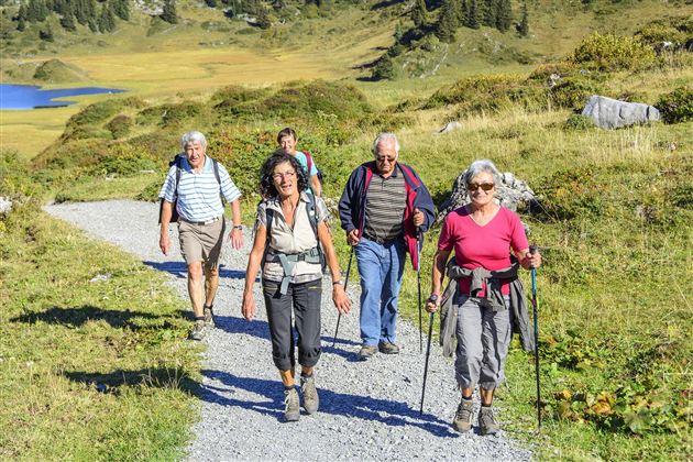 A group of five people is hiking on a gravel path through a picturesque landscape. In the background, green hills and a blue lake can be seen.