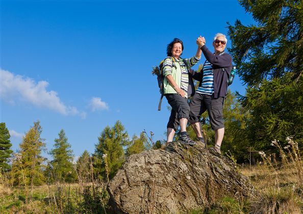 Two cheerful people are standing on a large stone outdoors. The sky is blue and there are trees in the background.