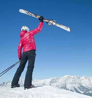 A person in a red ski jacket is lifting a pair of skis high. In the background, snow-covered mountains can be seen under a clear blue sky.