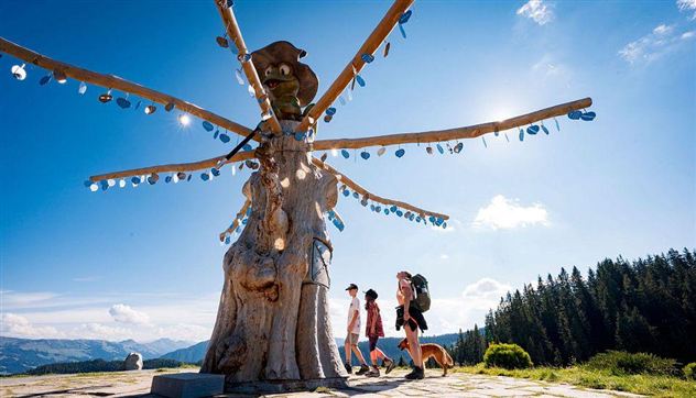An impressive wooden sculpture in nature with radiant decorative elements. Two people and a dog are walking along a path in front of the scene.
