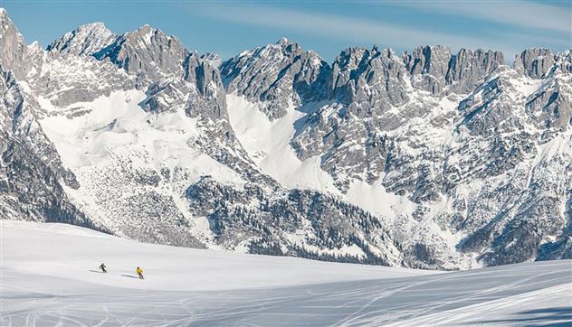A snowy landscape with majestic mountains in the background. Two skiers glide over the glistening snow.