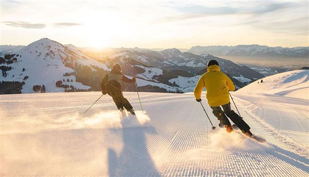 Zwei Skifahrer fahren bei Sonnenuntergang eine verschneite Piste hinunter. Im Hintergrund sind Berge und ein klarer Himmel zu sehen.