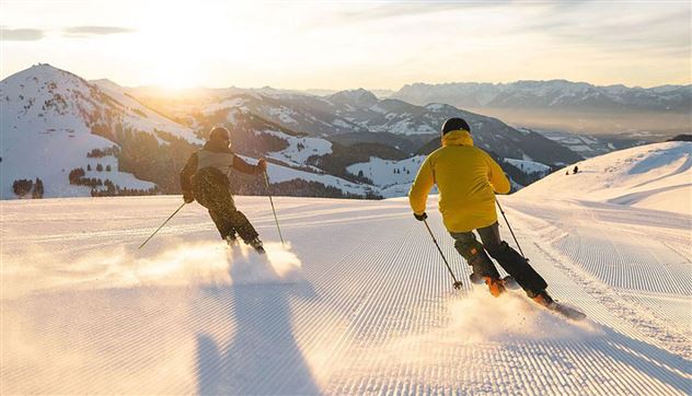 Two skiers are riding in the evening sun over a snow-covered slope. The mountains in the background provide a picturesque backdrop.