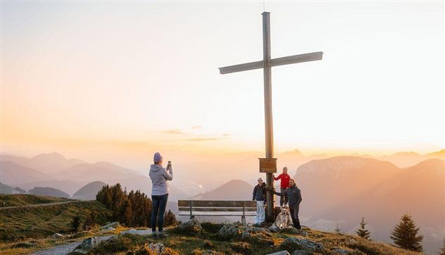 A group of people gathers around a large cross on a mountain. In the background, a picturesque mountain landscape can be seen at sunset.