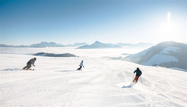 Three skiers enjoy a sunny descent on freshly groomed slopes. In the background, snow-covered mountains and a clear blue sky can be seen.