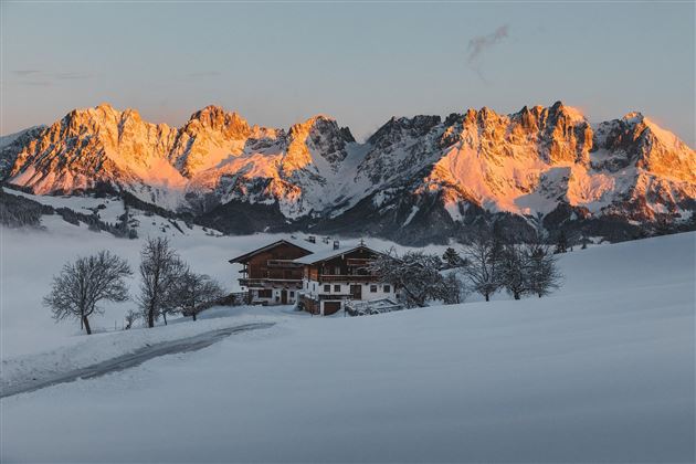 A picturesque winter landscape with snow-covered mountains and a traditional wooden house. The sun rays illuminate the mountain peaks in orange.