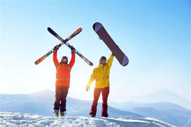 Two skiers are standing on a snowy peak, holding their skis and a snowboard up in the air. The sky is clear and blue, and the mountain landscape is visible in the background.