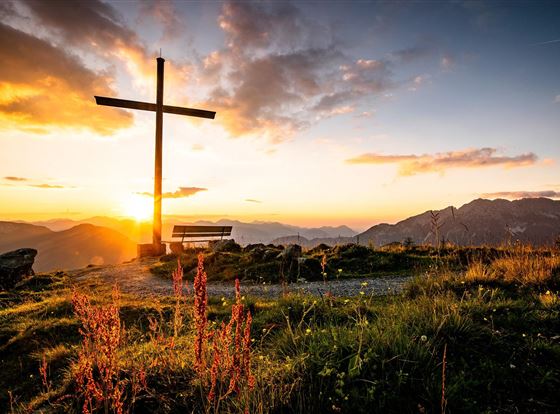 Ein großes Kreuz steht auf einem Hügel bei Sonnenuntergang. Die Umgebung ist wunderbar mit Bergen und Wiesen zu sehen.