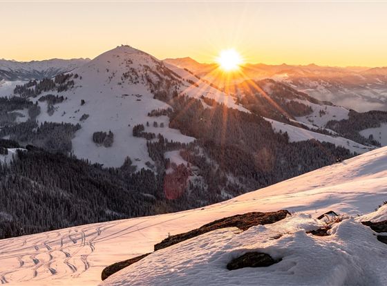 Eine verschneite Berglandschaft bei Sonnenaufgang. Die Sonne strahlt über die Berge und den Winterwald.