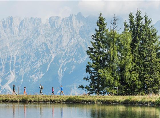 Eine Gruppe von Menschen wandert entlang eines ruhigen Sees, umgeben von hohen Bergen und Bäumen. Der Himmel ist klar und die Landschaft ist malerisch.