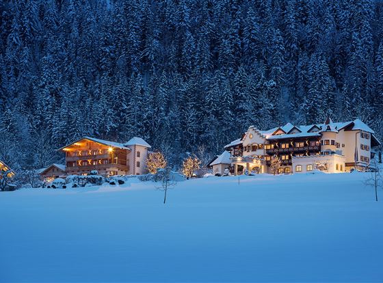 Eine winterliche Landschaft mit schneebedeckten Hügeln. Gemütliche Häuser leuchten sanft im Dämmerlicht.