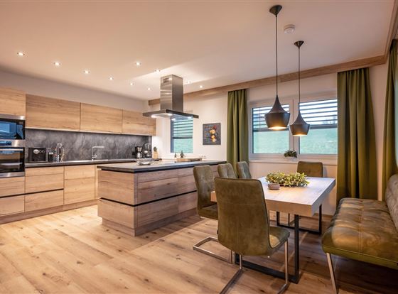Een moderne keuken met houten planken en een grote eettafel. De open sfeer wordt benadrukt door de elegante lampen en de groene gordijnen.