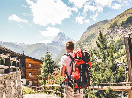 Ein Wanderer mit einem Rucksack steht vor einer beeindruckenden Berglandschaft. Im Hintergrund ist der Matterhorn sichtbar, umgeben von grünen Hügeln und klarer Blauen Himmel.