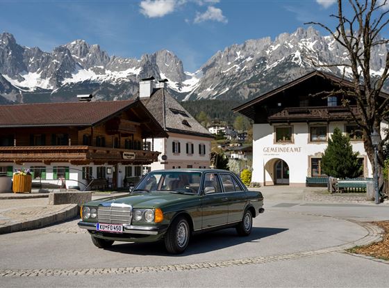 Ein alter Mercedes-Benz fährt durch ein malerisches Dorf mit traditioneller Architektur. Im Hintergrund ragen majestätische Berge unter einem klaren Himmel empor.