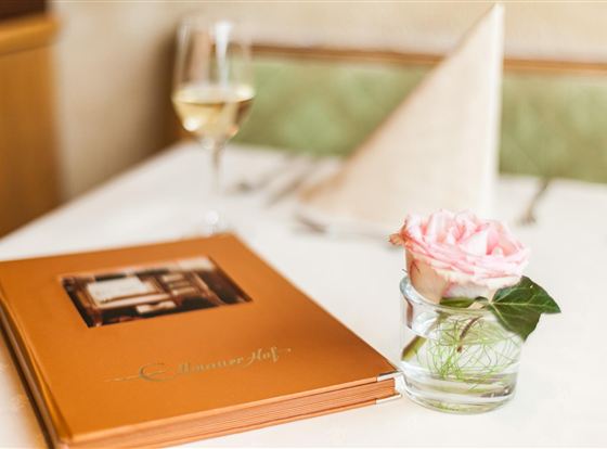 Ein eleganter Tisch mit einer Speisekarte und einer Rose in einer Glasvase. Im Hintergrund steht ein Glas Wein und ein gefaltetes Tuch auf dem Tisch.