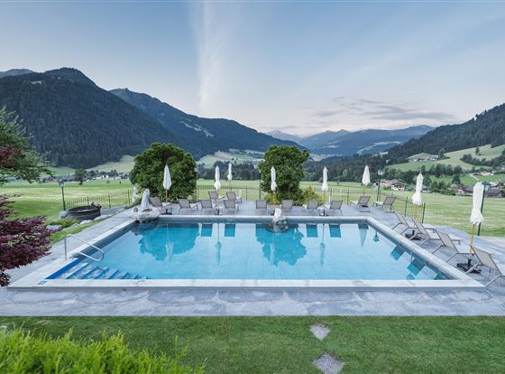 Ein schöner Pool mit Liegestühlen, umgeben von grünen Wiesen und Bergen. Der Himmel ist klar und zeigt sanfte Farben des Sonnenuntergangs.