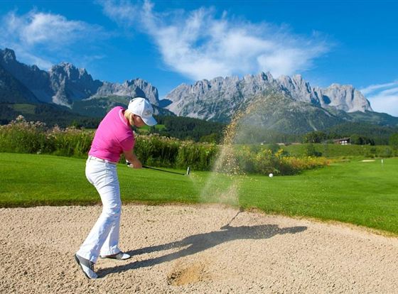 Ein Golfer schlägt aus einem Bunker auf einem schönen Golfplatz mit beeindruckenden Bergen im Hintergrund. Der Himmel ist klar und blau, die Landschaft ist grün und einladend.