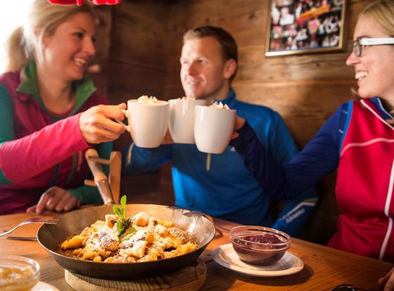 Drei Freunde stoßen mit Tassen an und genießen eine Mahlzeit in einer gemütlichen Umgebung. Auf dem Tisch liegt ein Teller mit köstlichen Leckereien.