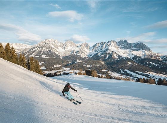 Ein Skifahrer fährt einen schneebedeckten Hang hinunter. Im Hintergrund sind majestätische Berge und ein klarer Himmel zu sehen.