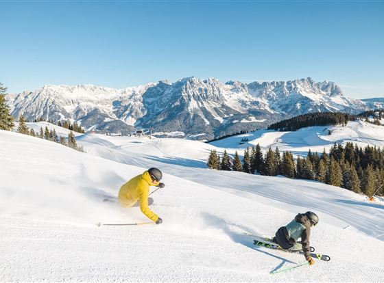 Zwei Skifahrer fahren durch frischen Schnee in den Alpen. Im Hintergrund sind majestätische Berggipfel zu sehen.