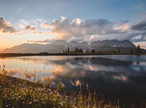 Ein malerischer See, umgeben von Bergen und blühenden Wiesen. Die untergehende Sonne spiegelt sich ruhig im Wasser.