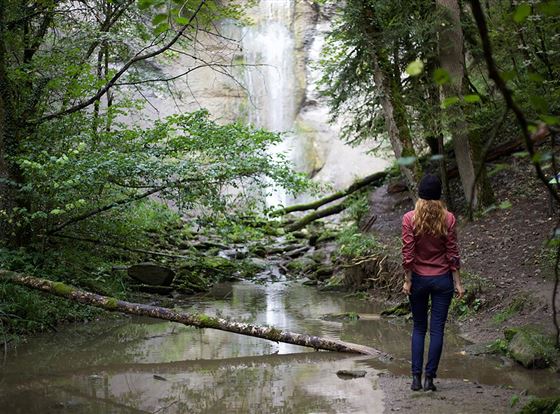 Eine Frau steht an einem kleinen Wasserfall in einem bewaldeten Gebiet. Der Wald ist grün und üppig, mit einem klaren Wasserbecken im Vordergrund.