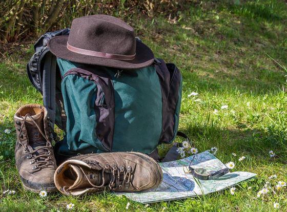 Ein Rucksack steht auf einer Wiese, neben einem Paar Wanderschuhe und einer Karte. Ein Hut liegt auf dem Rucksack und im Hintergrund sind frisches Gras und kleine Blumen zu sehen.