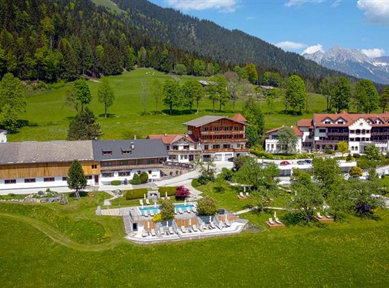 Eine malerische Landschaft mit einem Hotelkomplex, umgeben von Wäldern und Bergen. Im Vordergrund sind ein Poolbereich und grüne Wiesen zu sehen.