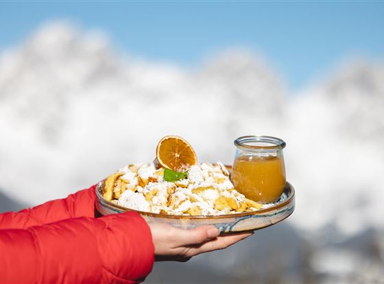 Ein Teller mit frischen, süßen Speisen, garniert mit einer getrockneten Orange und einem Glas Saft. Im Hintergrund sind schneebedeckte Berge sichtbar.