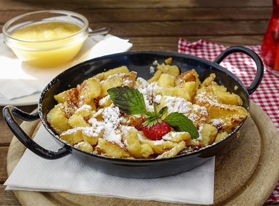 Ein köstlicher Kaiserschmarrn, serviert in einer Pfanne, mit Puderzucker, frischen Beeren und Minze. Dazu gibt es eine Schüssel Apfelmus.