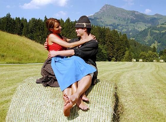 EinPaar sitzt auf einem Heuballen in einer grünen Wiese mit Bergen im Hintergrund. Sie lächeln sich an und genießen die schöne Landschaft.