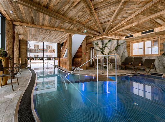 Ein eleganter Innenpool in einem Holzgebäude mit großen Fenstern. Komfortable Liegen und natürliche Elemente schaffen eine entspannende Atmosphäre.