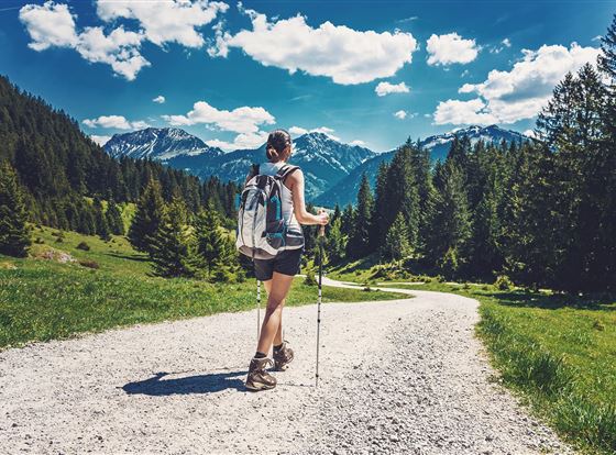 Eine Person wandert auf einem schotternden Weg durch eine grüne Landschaft. Im Hintergrund sind Berge und ein blauer Himmel mit einigen Wolken zu sehen.