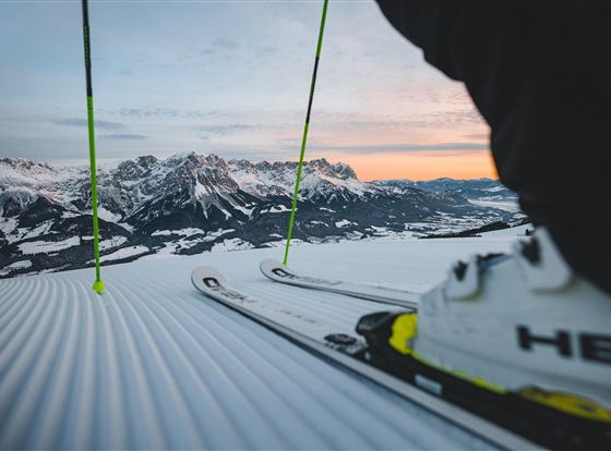 Eine winterliche Berglandschaft mit schneebedeckten Gipfeln. Im Vordergrund sind Skier und Skistöcke zu sehen, während die Sonne aufgeht.