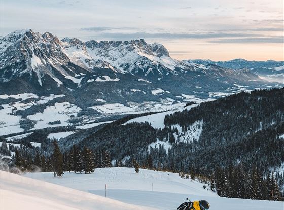 Ein Skifahrer in einem gelben Anzug fährt die verschneite Piste hinunter. Im Hintergrund sind majestätische Berge und eine ruhige Winterlandschaft zu sehen.