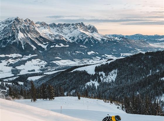 Ein Skifahrer in gelber Kleidung fährt eine Piste hinunter. Die Berge und das verschneite Tal sind im Hintergrund sichtbar.