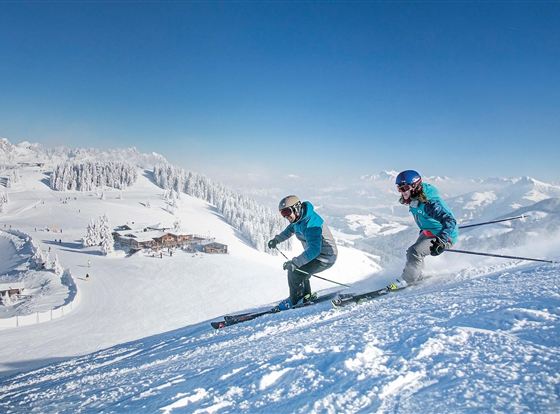 Zwei Skifahrer fahren die verschneite Piste hinunter. Im Hintergrund sind schneebedeckte Berge und ein blauer Himmel zu sehen.