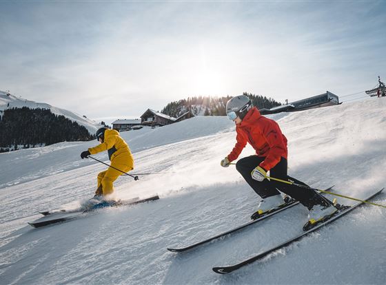 Zwei Skifahrer fahren auf einer verschneiten Piste. Die Sonne scheint und es gibt Schnee im Hintergrund.