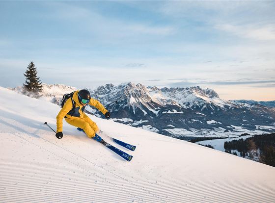 Ein Skifahrer in gelber Ausrüstung fährt auf frischem Schnee. Im Hintergrund sind beeindruckende Berge und ein klarer Himmel zu sehen.