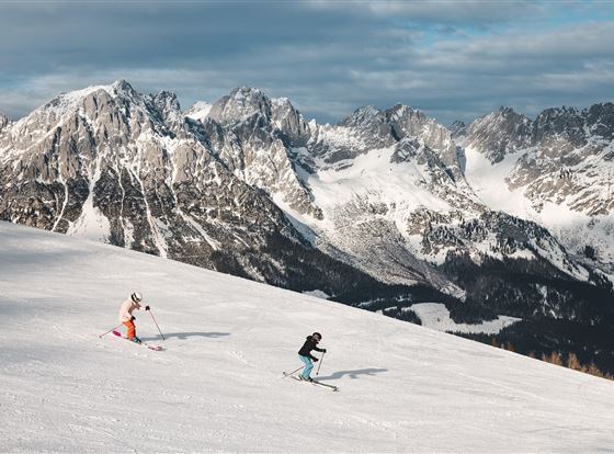 Zwei Skifahrer fahren die verschneite Piste hinunter. Im Hintergrund sind imposante Berge unter einem grauen Himmel zu sehen.