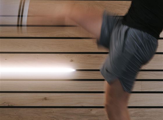 Ein Mann führt einen dynamischen Kick aus. Im Hintergrund sind holzverkleidete Wände und eine moderne Beleuchtung zu sehen.
